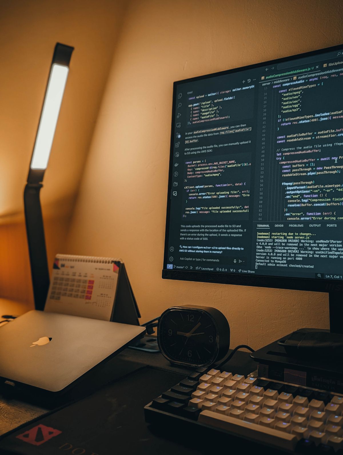 A working desk at night — code editor open on the monitor, lamp on